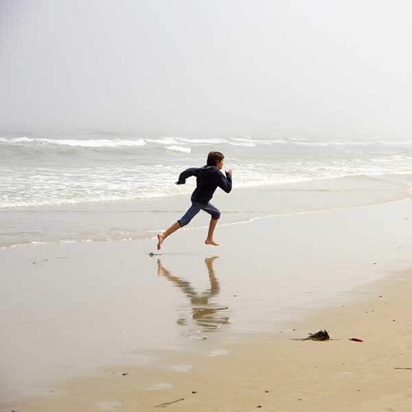 A young woman wearing jeans and a hoodie running on the shoreline of a cold beach.