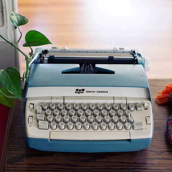A vintage typewriter with raised buttons sitting on a wooden table with a vine growing from its side.